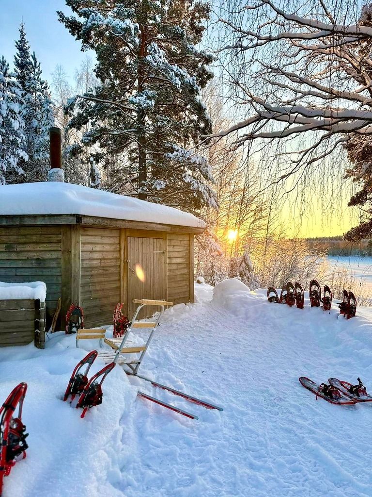 Besneeuwde scène met een houten sauna, sneeuwschoenen en een zonsopgang boven een bevroren meer.