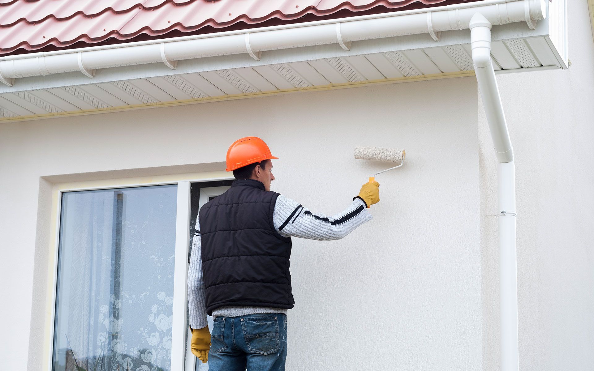Painter in orange hard hat, painting white house exterior with a roller.