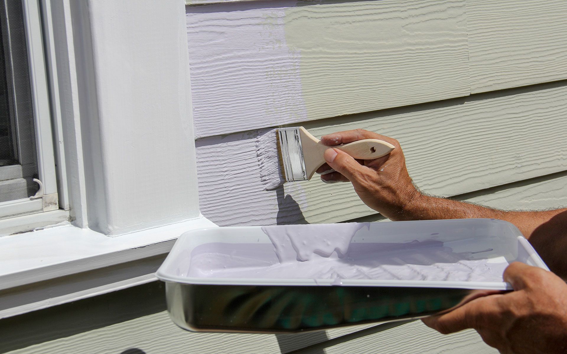 Person painting exterior siding with a brush and paint tray; white trim and a window are visible.