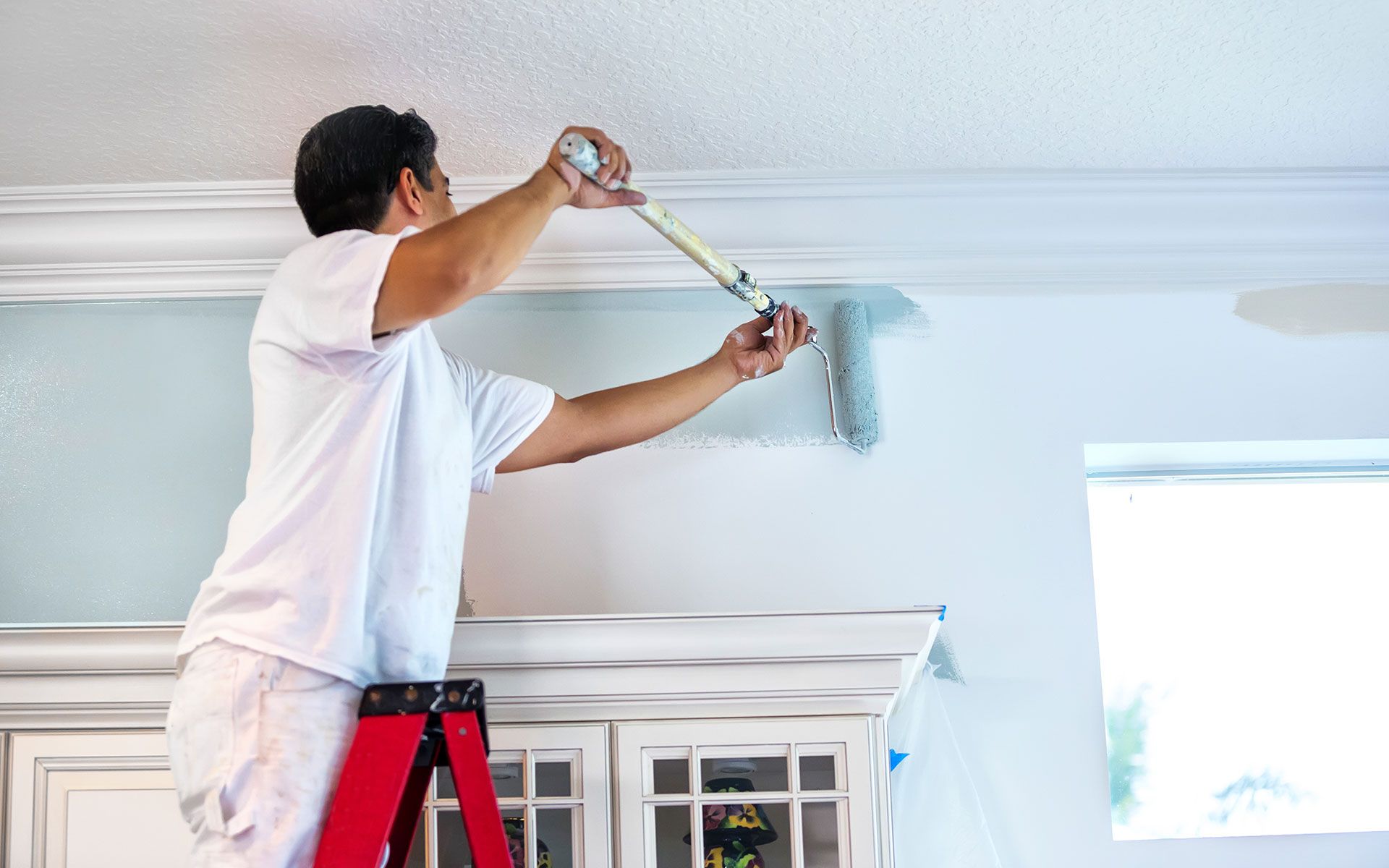 Man on a stepladder painting a wall blue with a roller, next to a white cabinet.