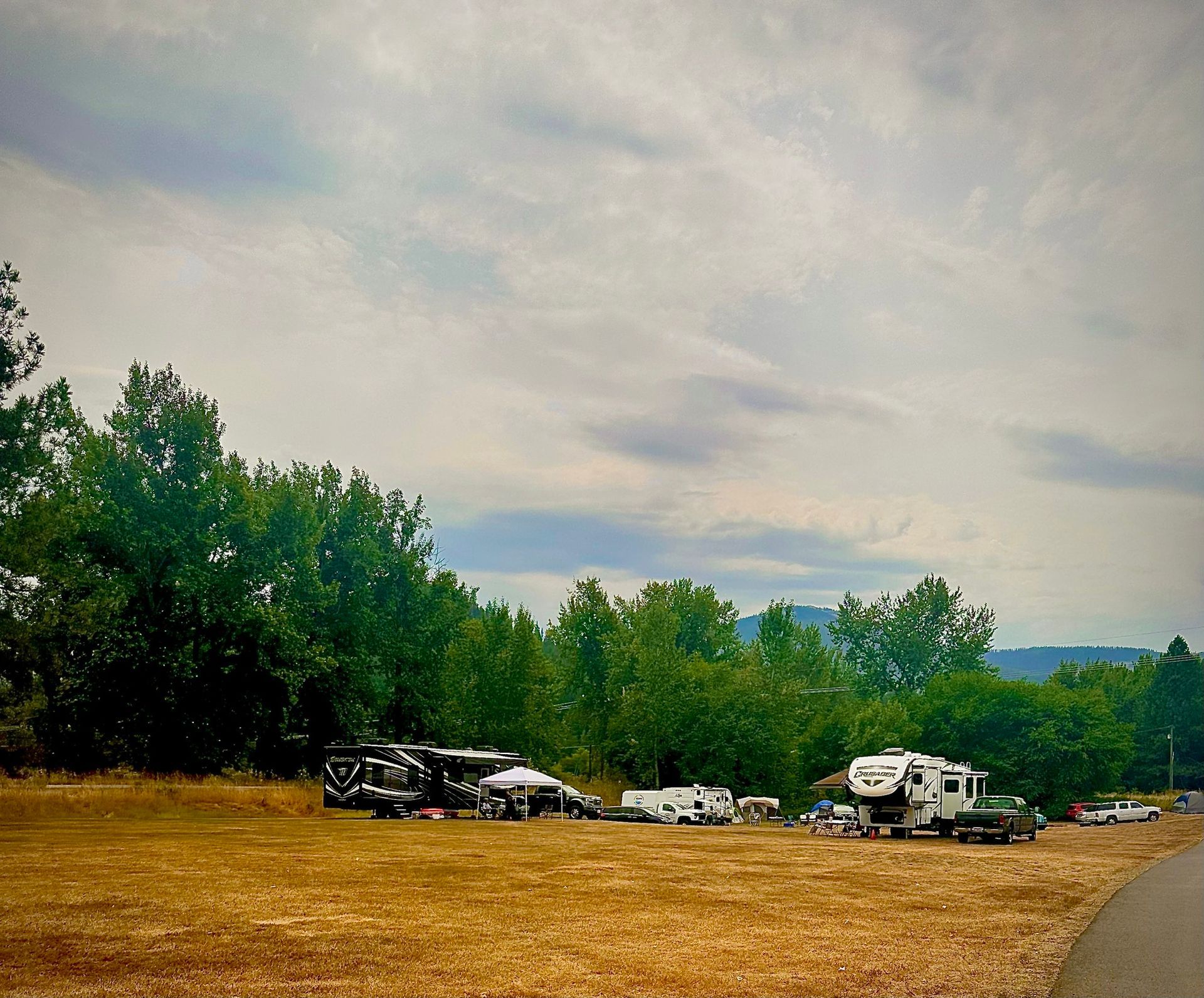 Campground with RVs parked on dry grass, trees in the background, under a cloudy sky.