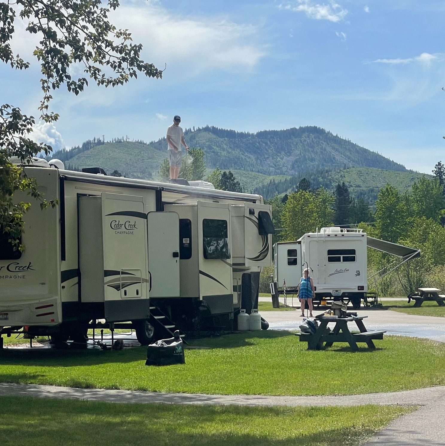 Person on RV roof, washing it, campsite with mountain backdrop.