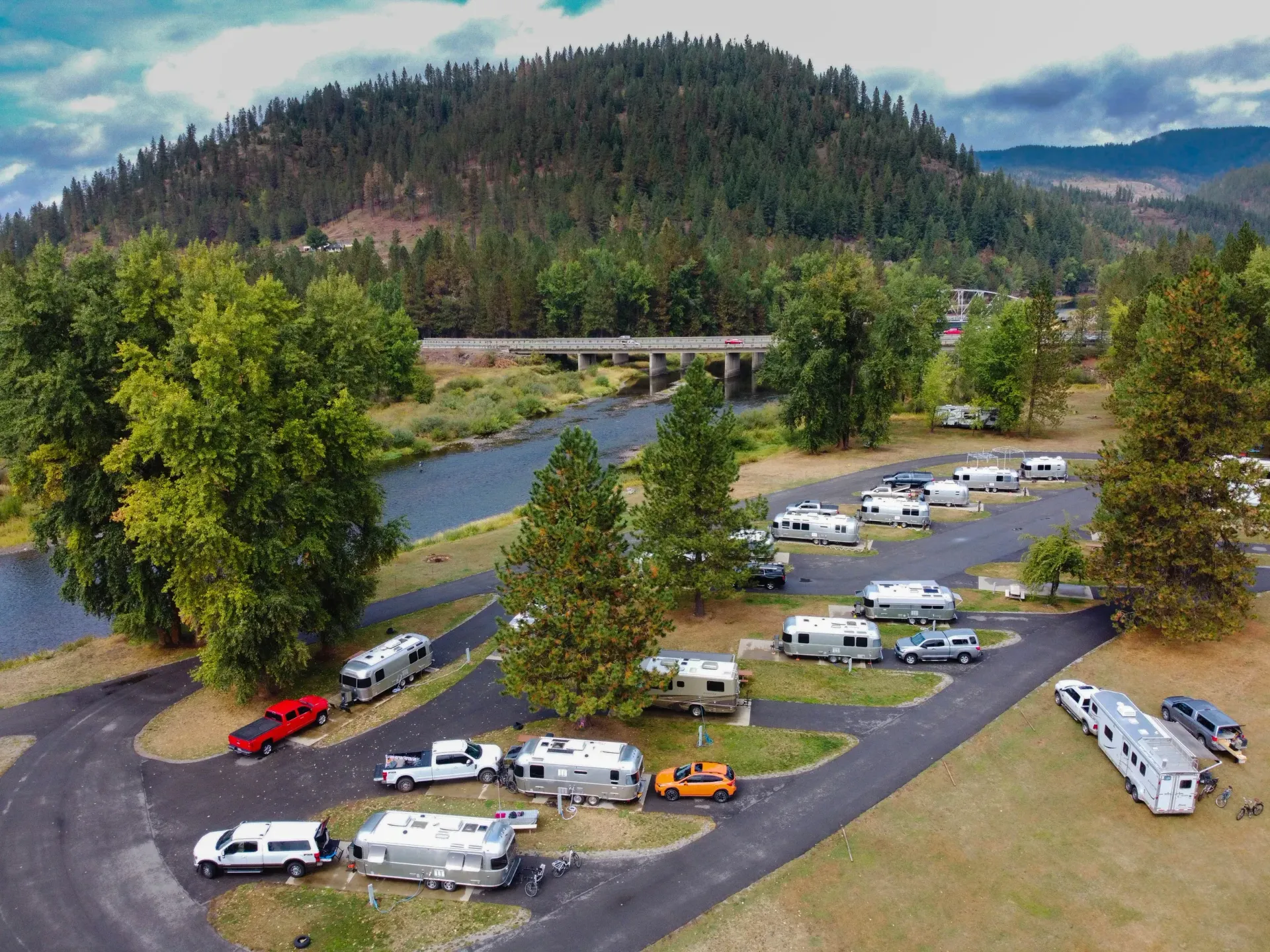 RV park with many RVs parked on paved lots near a river and a forested hill.
