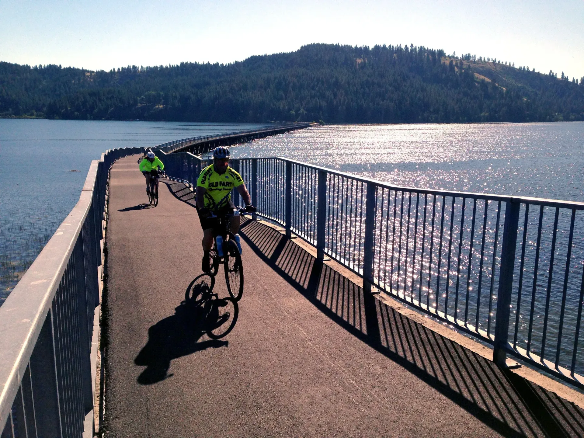 Two cyclists ride bikes on a path over a lake, sunny day, blue water, green trees in background.