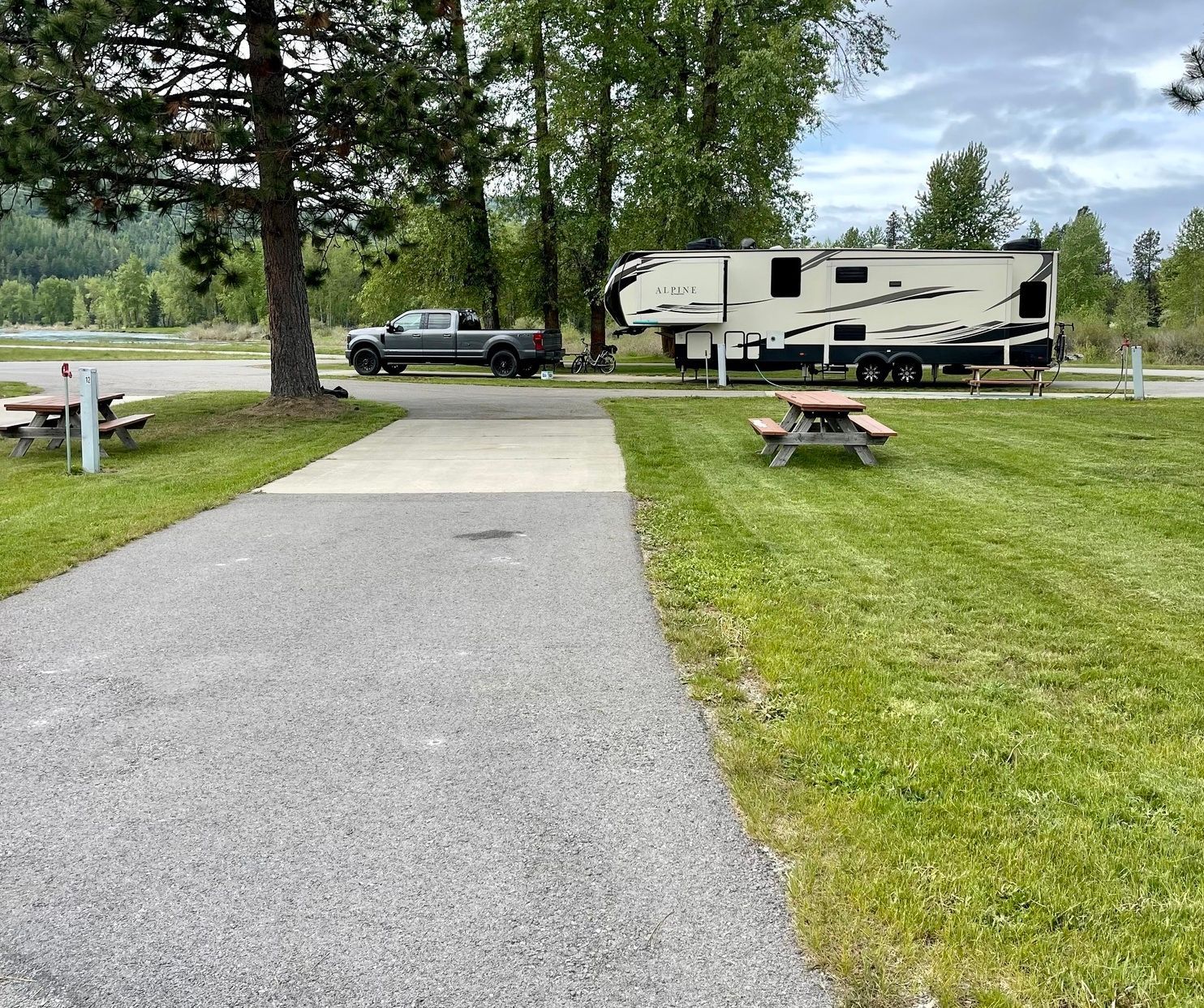 Campsite with RV and truck parked on gravel pad and grass, picnic tables, trees, and overcast sky in the background.