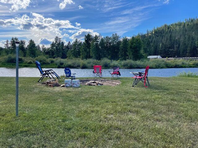 Camp chairs around a fire pit by a river. Green grass, trees, and cloudy blue sky.