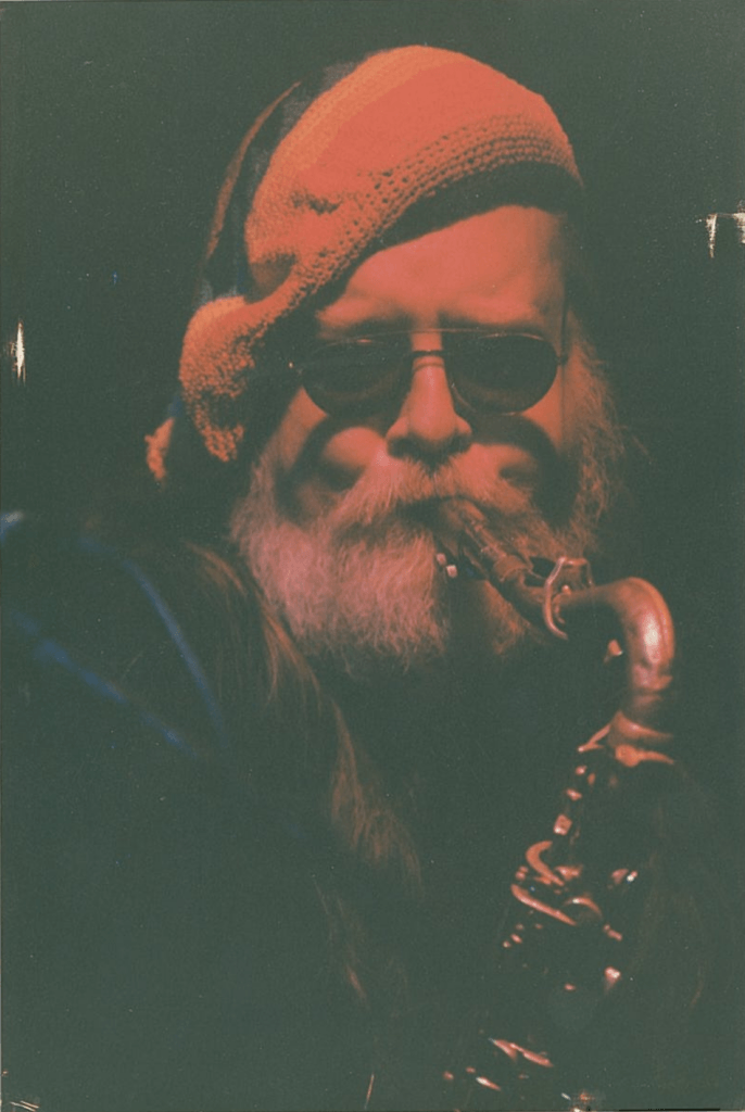 Man with sunglasses and a white beard playing a saxophone, wearing a knitted hat.