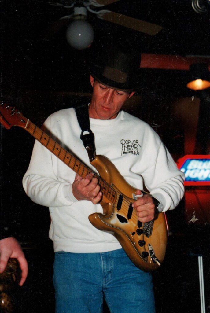 Man in hat and sweatshirt playing a sunburst electric guitar in a dimly lit bar.