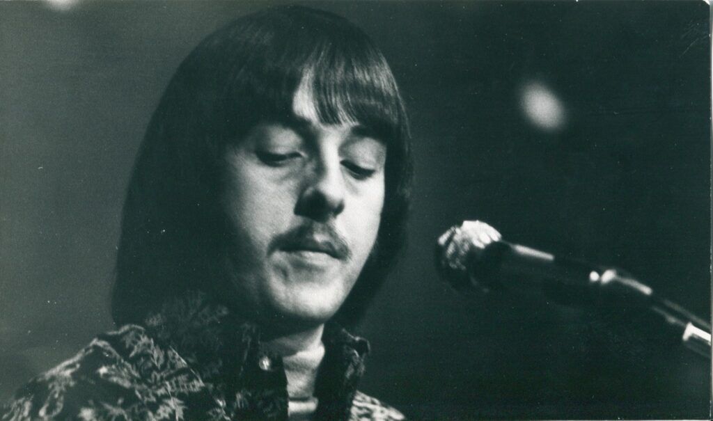 Man with dark hair and mustache singing into a microphone, eyes closed.