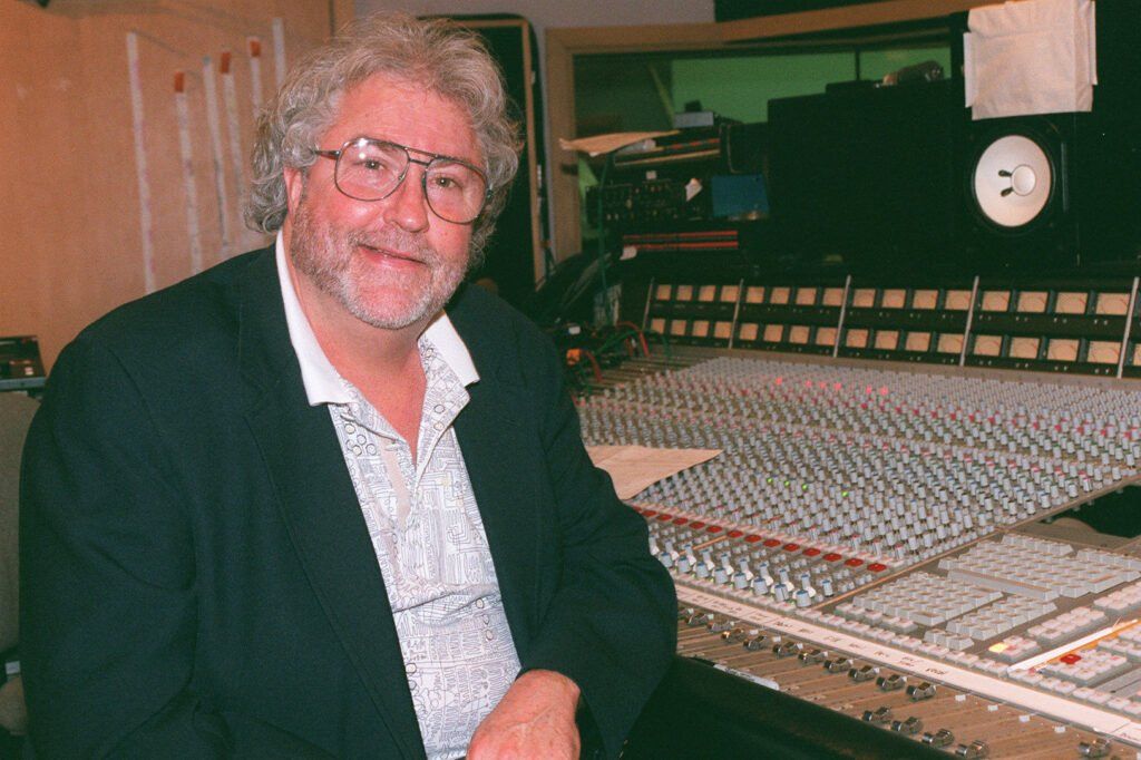 Man with glasses sits at a sound mixing console. He smiles in a recording studio setting.