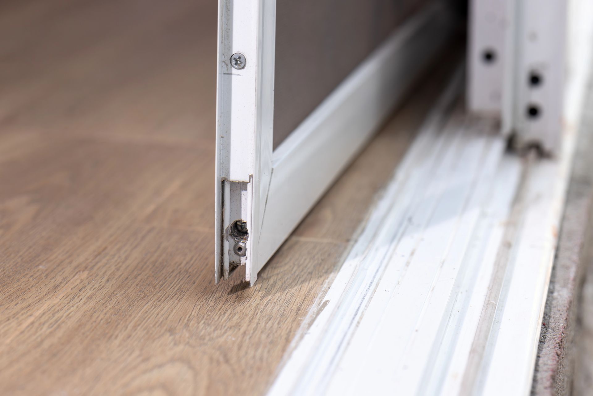 Il bordo inferiore di una porta scorrevole a zanzariera bianca è posizionato sopra un binario su un pavimento in legno chiaro.