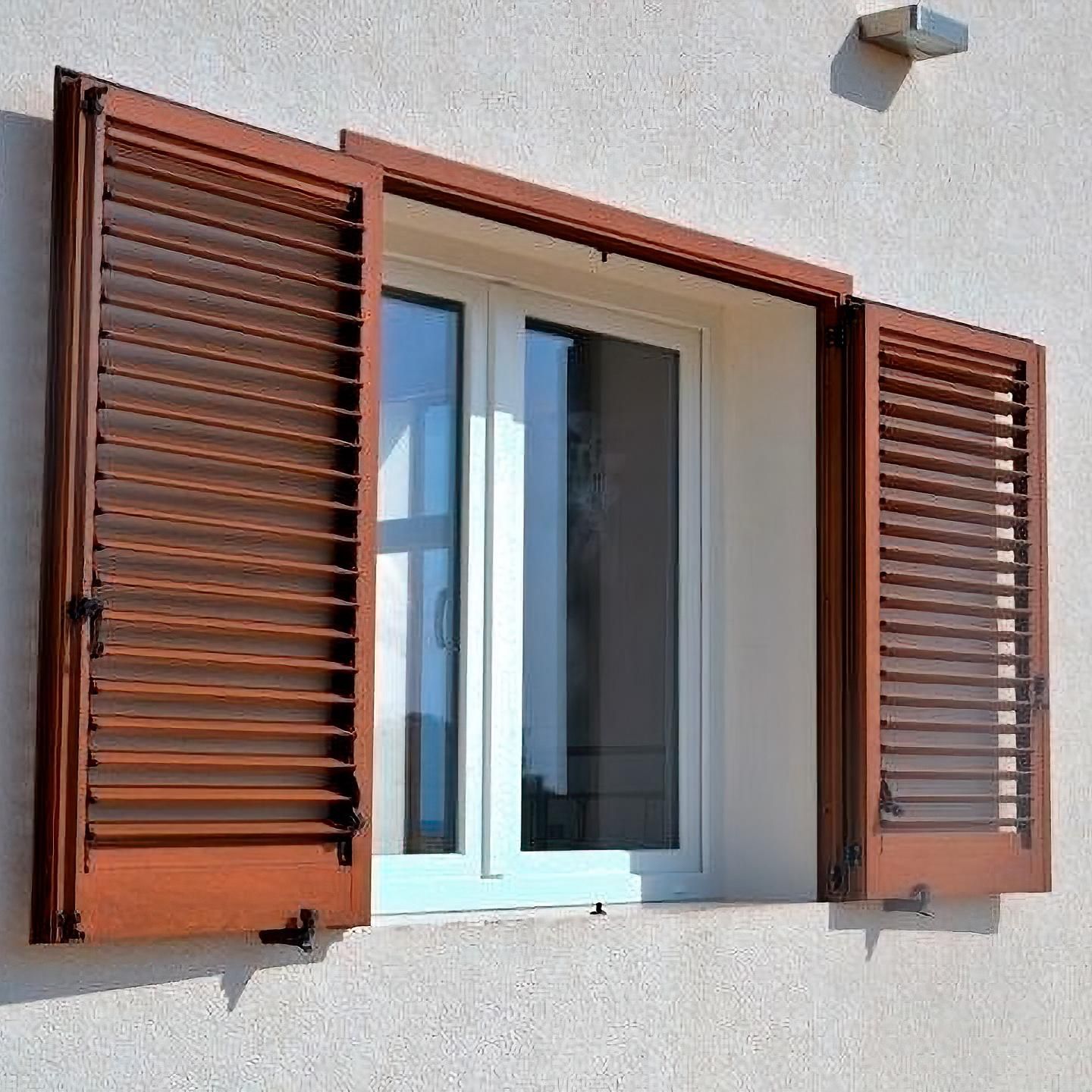 Una finestra bianca con persiane a lamelle di legno marrone aperte, incastonata in una parete di stucco beige.