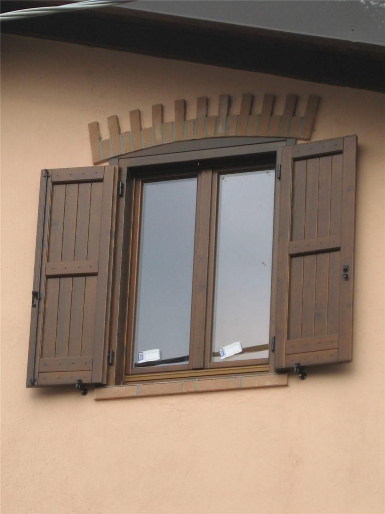 Una finestra marrone con persiane di legno aperte e muratura decorativa sopra, incastonata in una parete beige.