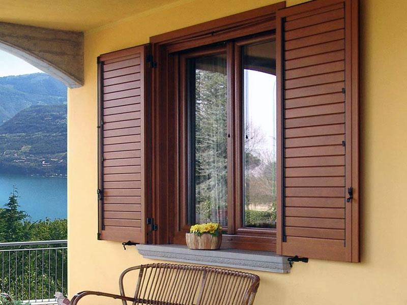 Una finestra con persiane di legno aperte su una parete esterna gialla, con vista su un lago e sulle montagne.