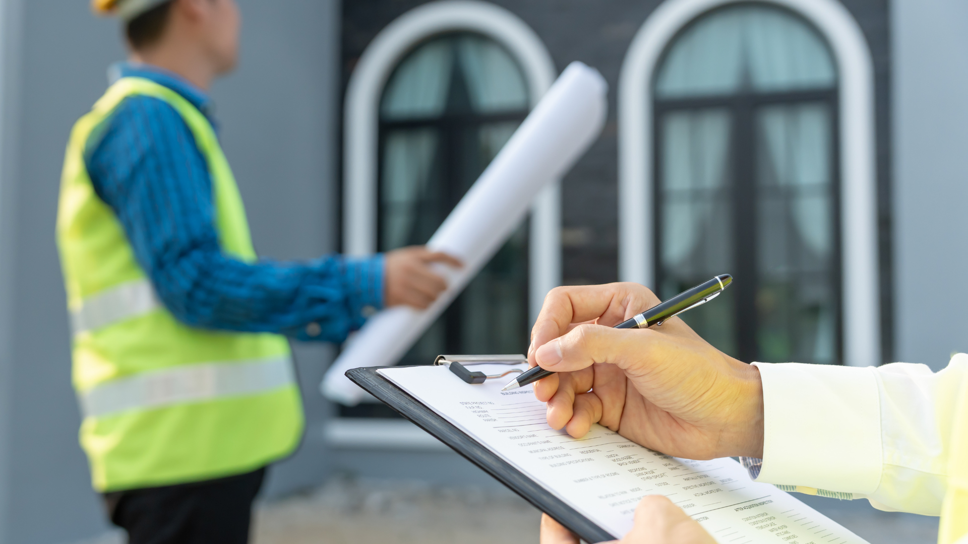 A person writes on a clipboard while a construction worker in a high-visibility vest holds blueprints in the background.