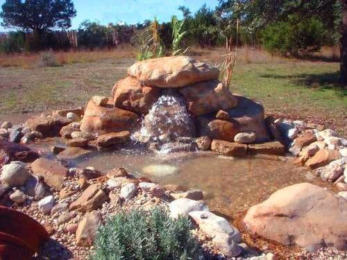 A small waterfall is surrounded by rocks in a field.