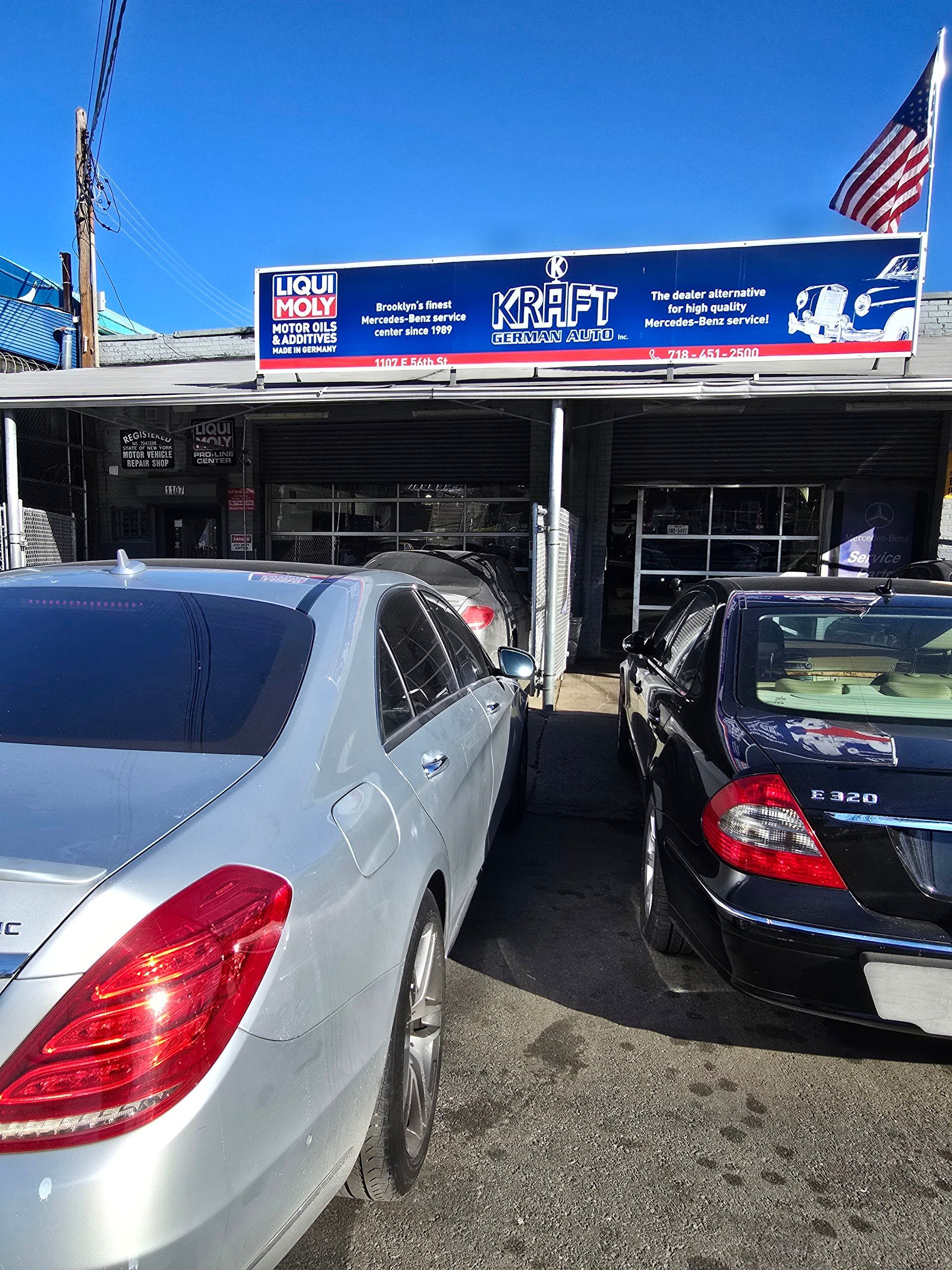 Silver car parked outside an auto repair shop with the American flag. Black car next to it. | Kraft German Auto