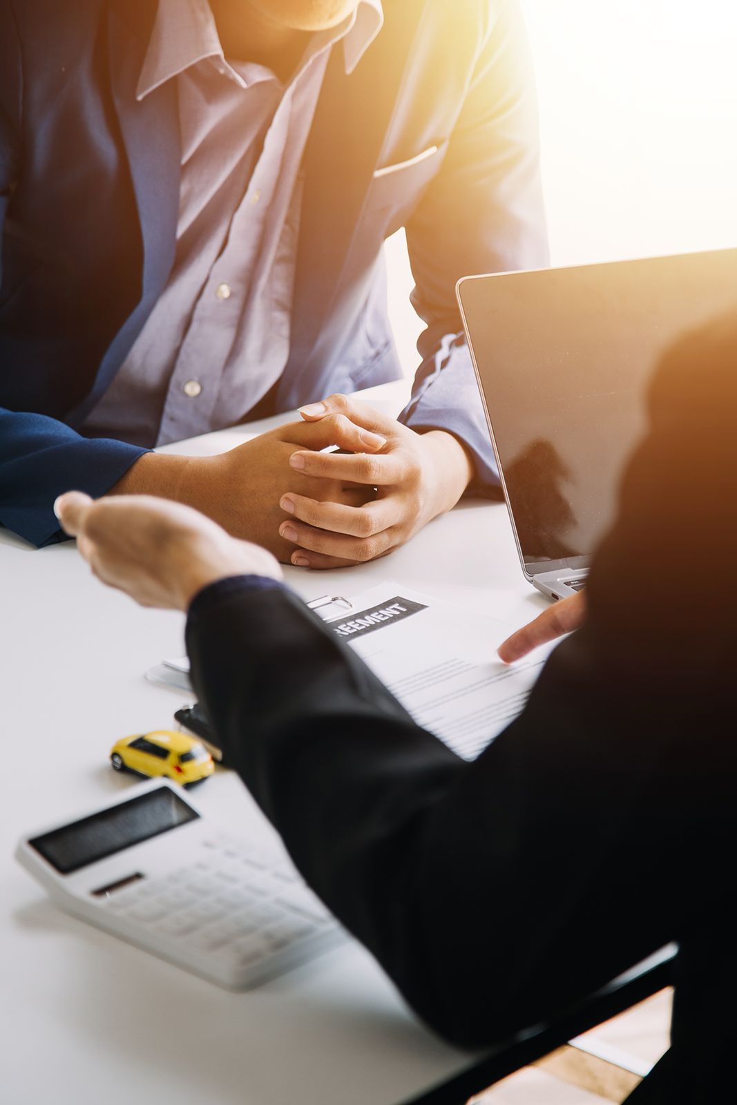 Dos personas, una con traje señala documentos, la otra tiene las manos entrelazadas, un modelo de coche y una calculadora sobre la mesa.