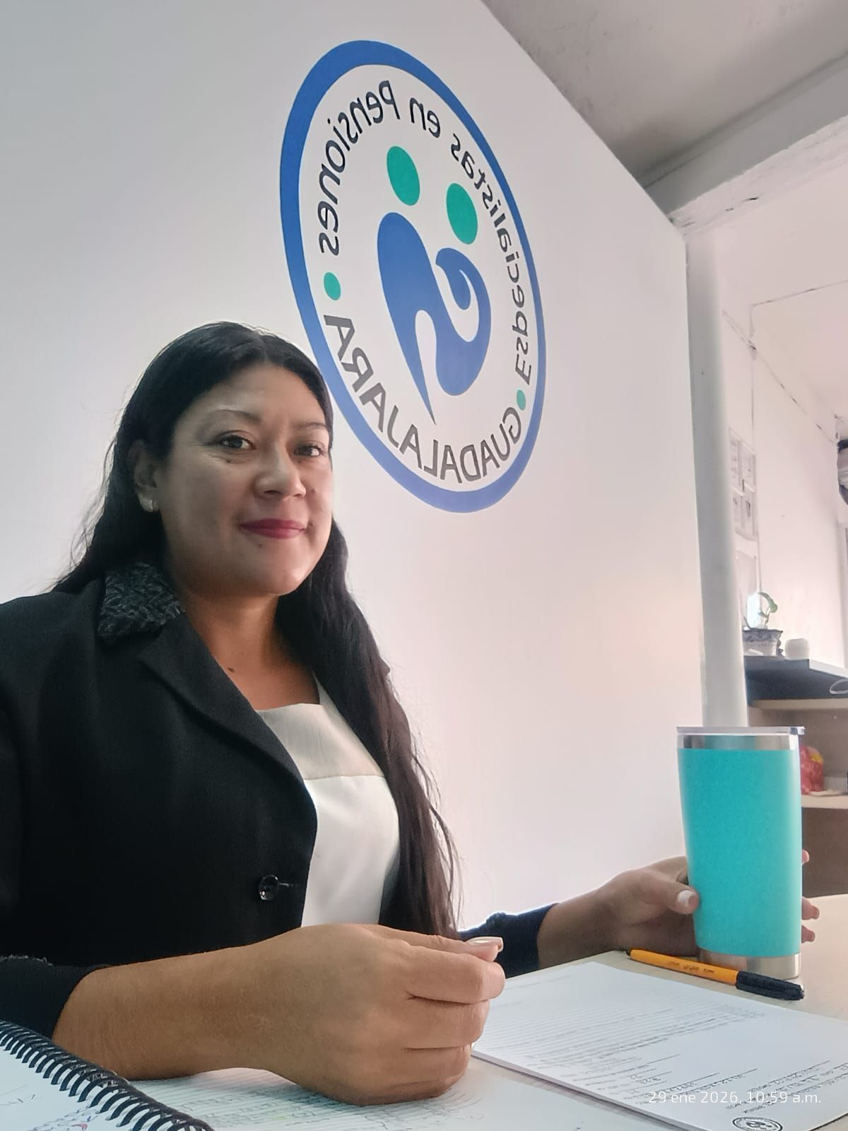 Mujer sosteniendo una taza turquesa, sentada en un escritorio, con un logotipo en la pared detrás de ella.