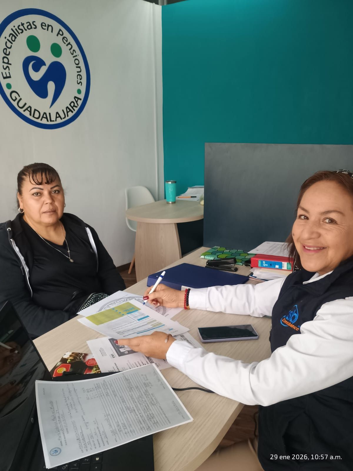 Dos mujeres en una mesa; una escribe, la otra está sentada. Oficina azul y blanca con el logo de 