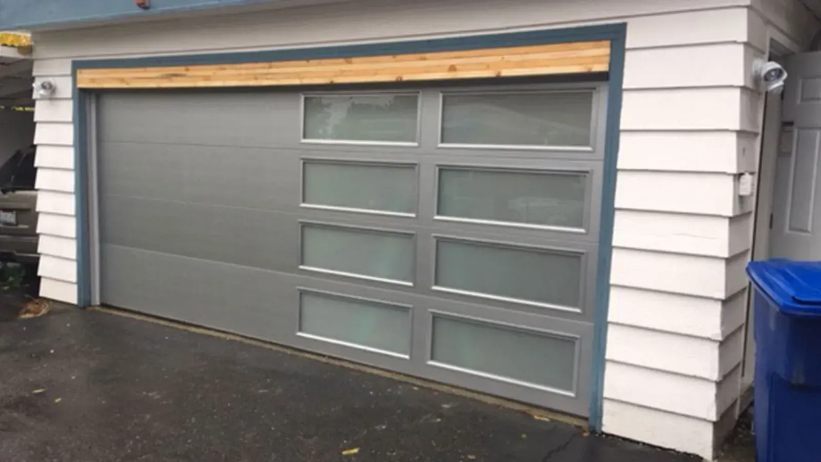 A modern gray garage door with a column of frosted glass panels on the right, installed on a white-sided building.