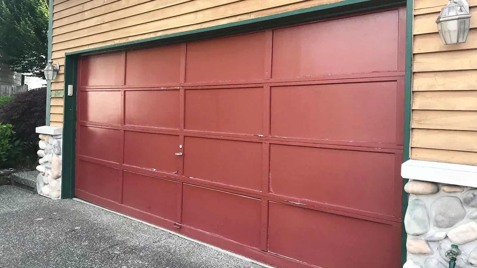 A red, rectangular, paneled garage door set in a home with wooden siding and stone-accented pillars.