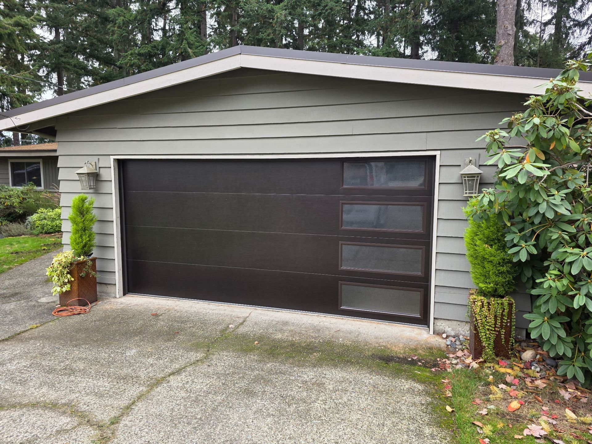 A dark brown modern garage door with vertical windows on the right, attached to a gray, horizontal-sided suburban house.