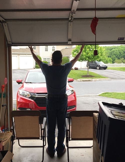 A person stands with arms raised, holding up a partially open garage door, facing a red car in a suburban driveway.