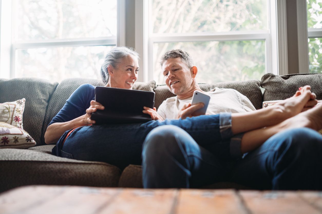 Two people sit together on a sofa, looking at a tablet and a smartphone in a brightly lit living room.