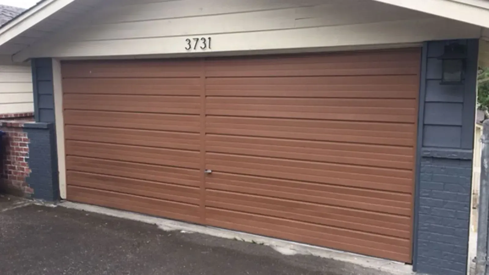 A brown garage door with horizontal panels, numbered 3731, flanked by grey brick columns under a beige house gable.