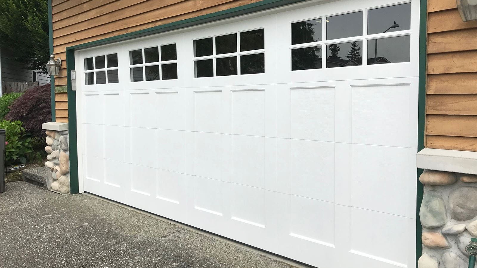 A white garage door with a row of window panes, set in a house with light brown siding and stone pillar accents.