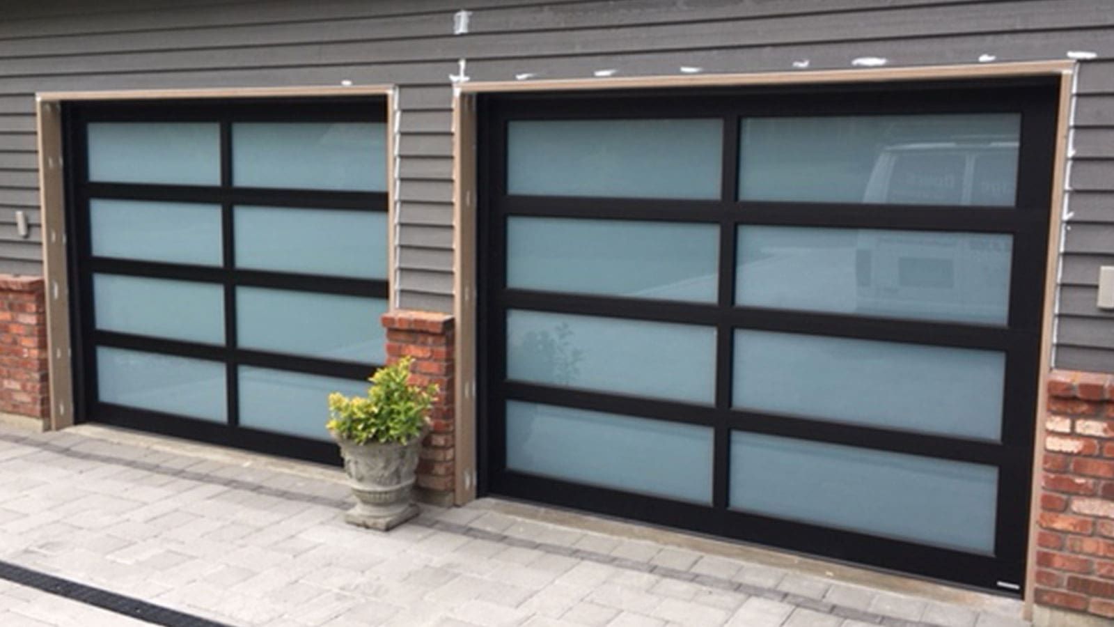 Two modern garage doors with black frames and frosted glass panels set in a grey house with brick accents.