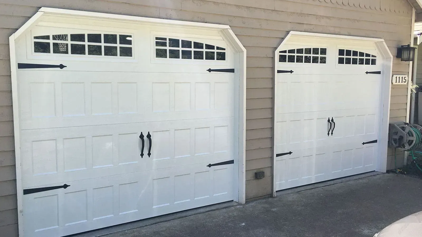 Two white garage doors with decorative black hinges, handles, and rectangular window panels installed on a house exterior.