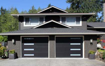 A dark-painted two-story house with two modern garage doors featuring horizontal window panels, set against greenery.