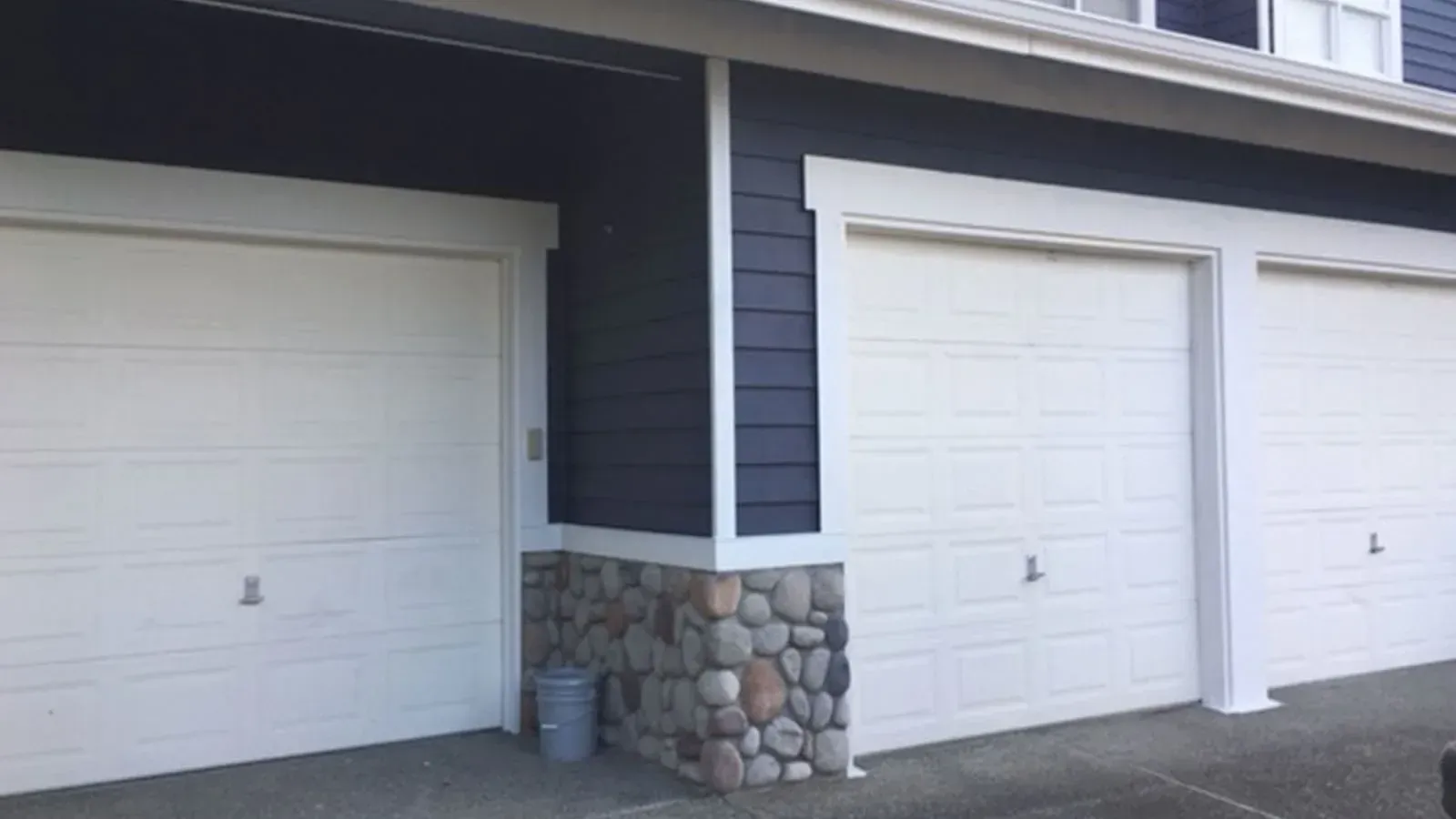 Three white residential garage doors set against dark blue siding with a stone-accented corner pillar.