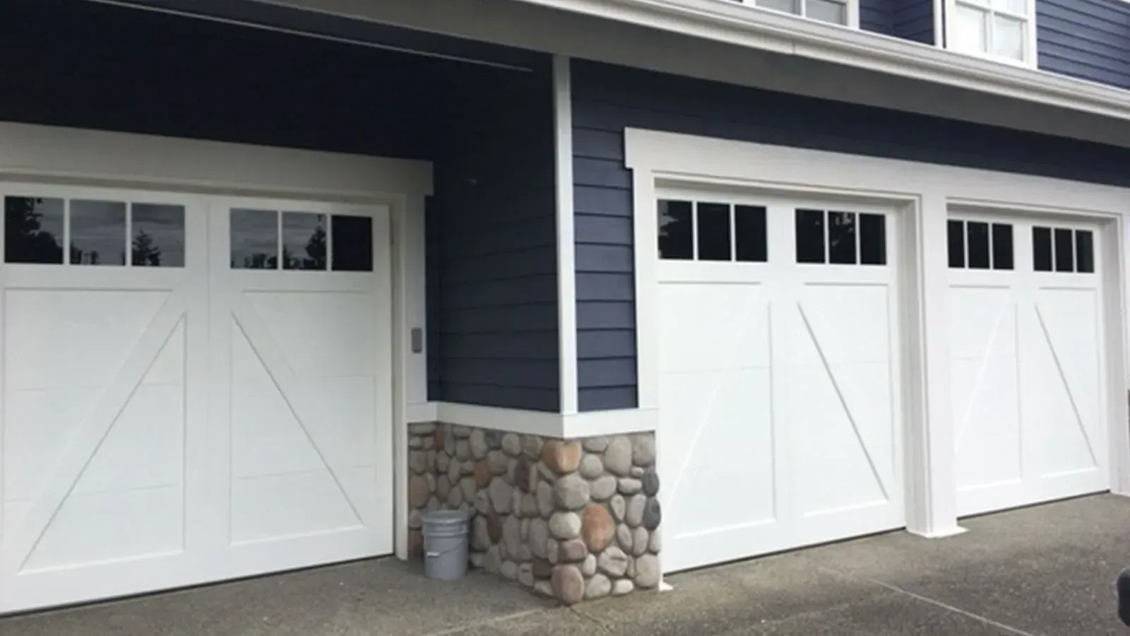 Three white carriage-style garage doors with windows on a dark blue house with stone accents.