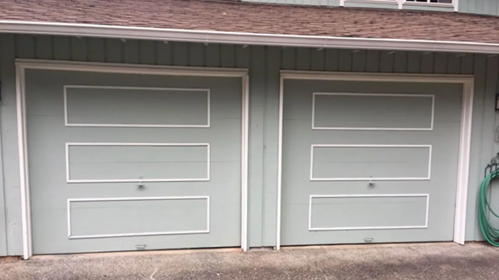 Two light green garage doors with three rectangular panels each, set in a wood-paneled wall under a shingled roof.