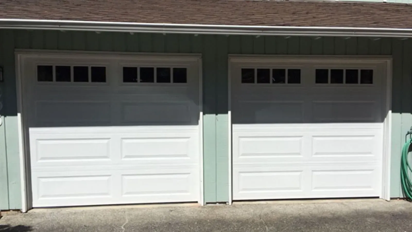 Two white garage doors with rectangular windows on a house with light green siding.