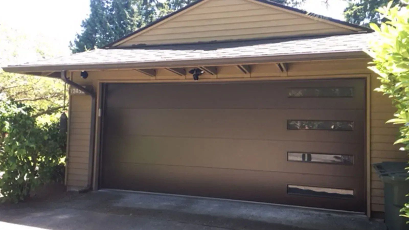 A modern brown garage door with a vertical row of four narrow windows on the right side, set in a beige-sided home.