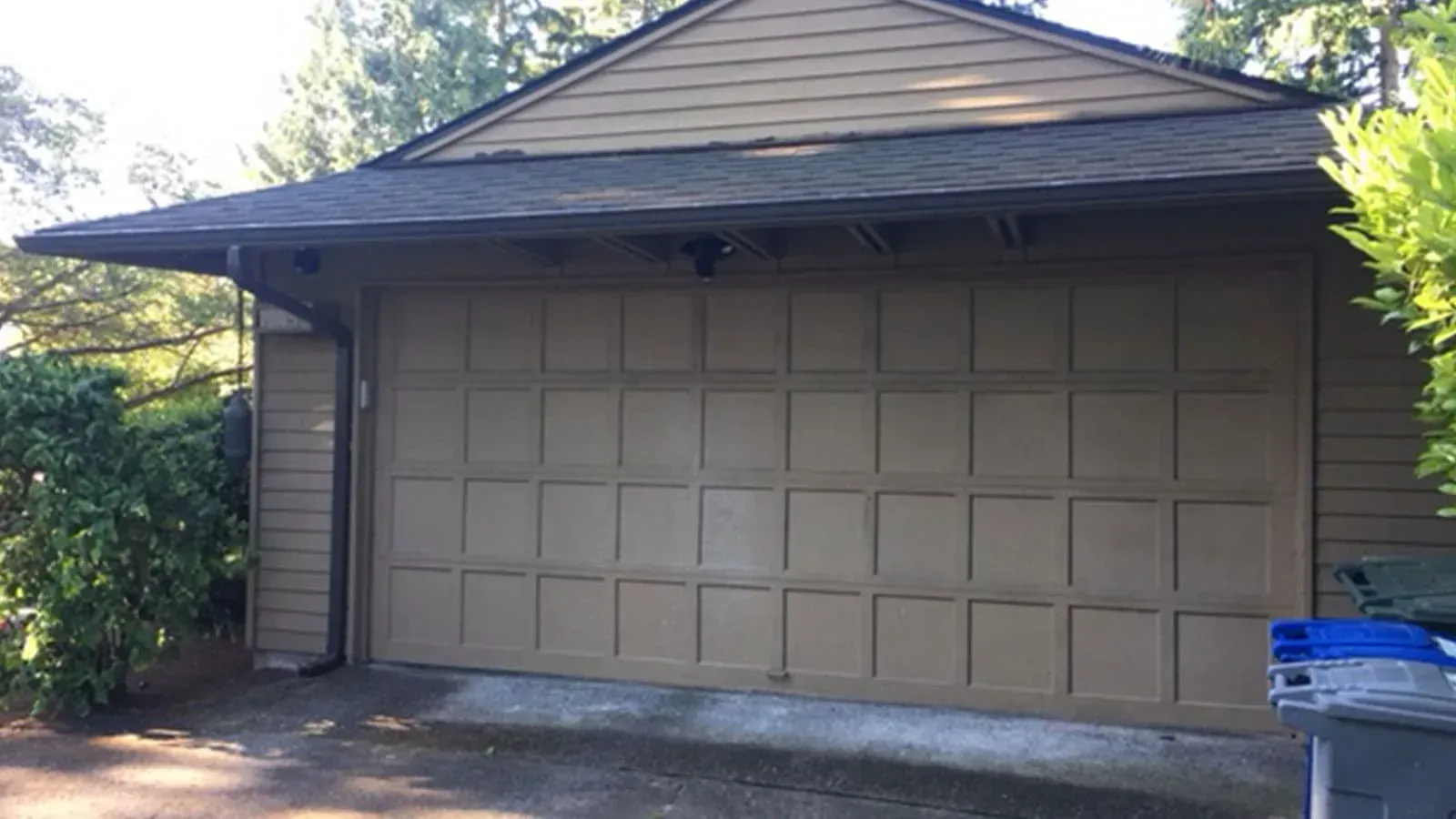 A tan, paneled garage door on a house with brown siding, a dark shingled roof, and an overhanging eave.