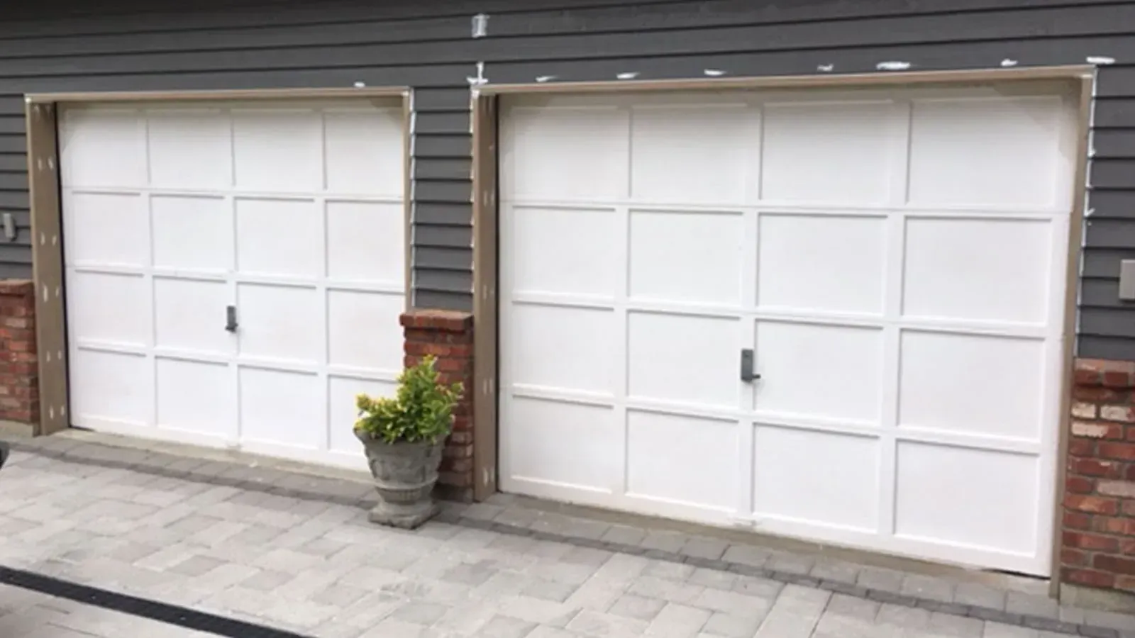 Two white garage doors with a grid panel design, installed in a gray siding wall with brick accents and a potted plant.