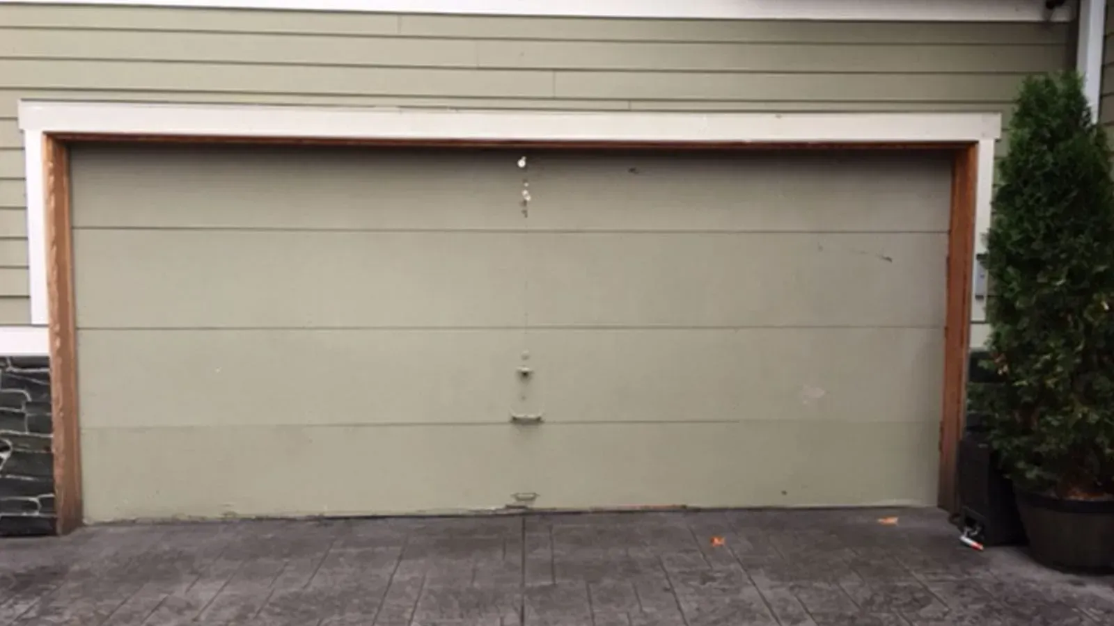 A closed, light olive-green garage door with horizontal panels, set in a house exterior with a paved driveway.