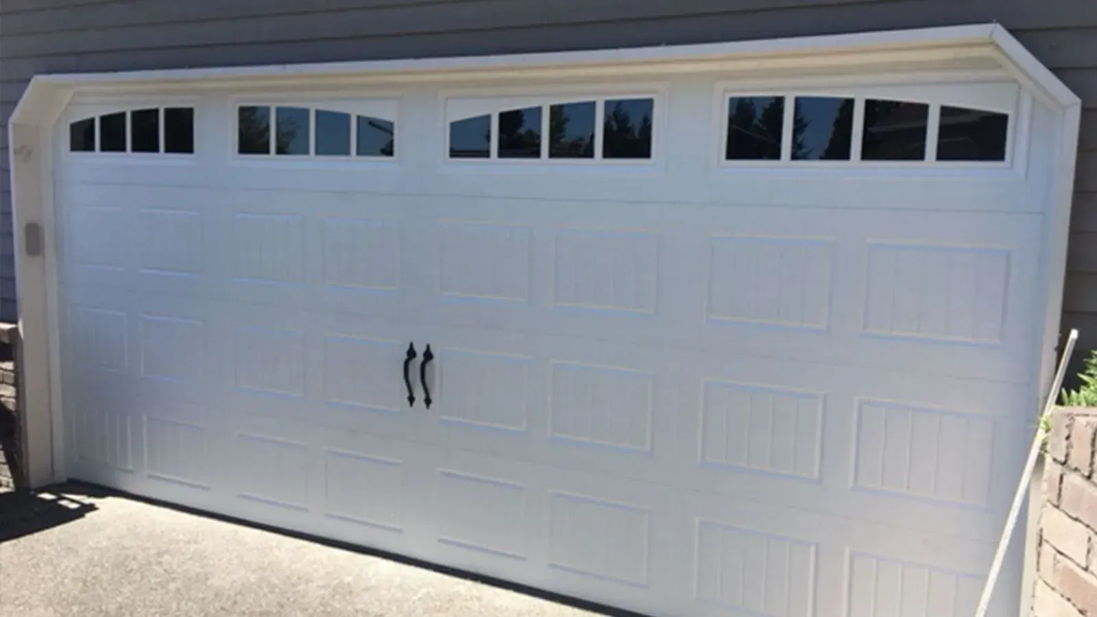 A white garage door with rectangular panels and three top windows with decorative inserts and two black handles.