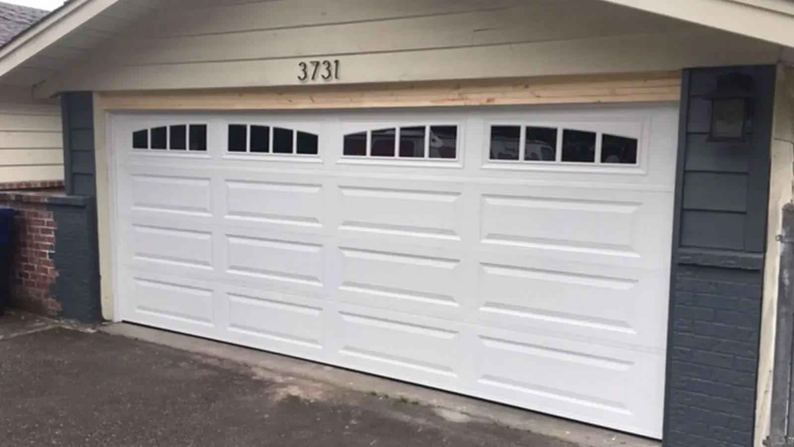 A white multi-panel residential garage door with arched, decorative top windows below a wooden header on a house exterior.