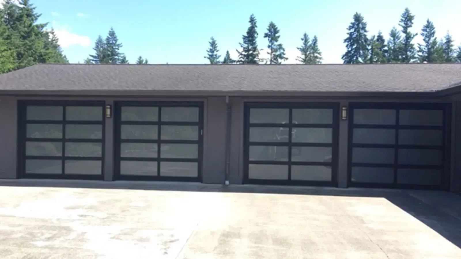 A gray garage with four modern, black-framed glass garage doors with frosted panes, set before a treeline.