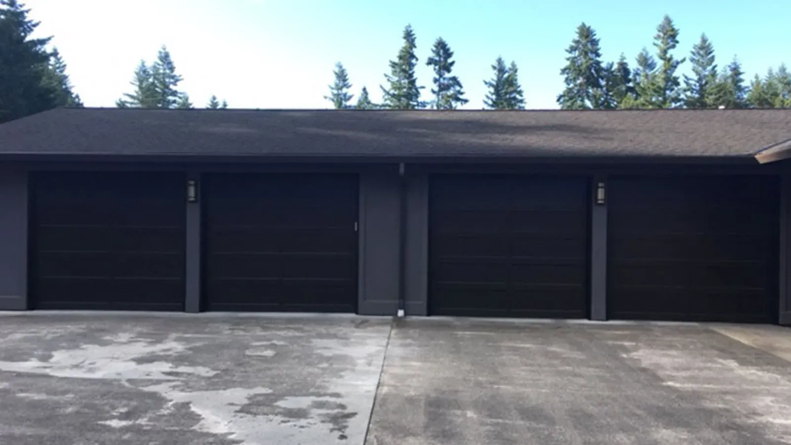 A dark grey, four-car garage building with matching doors under a shingled roof, set against a backdrop of pine trees.