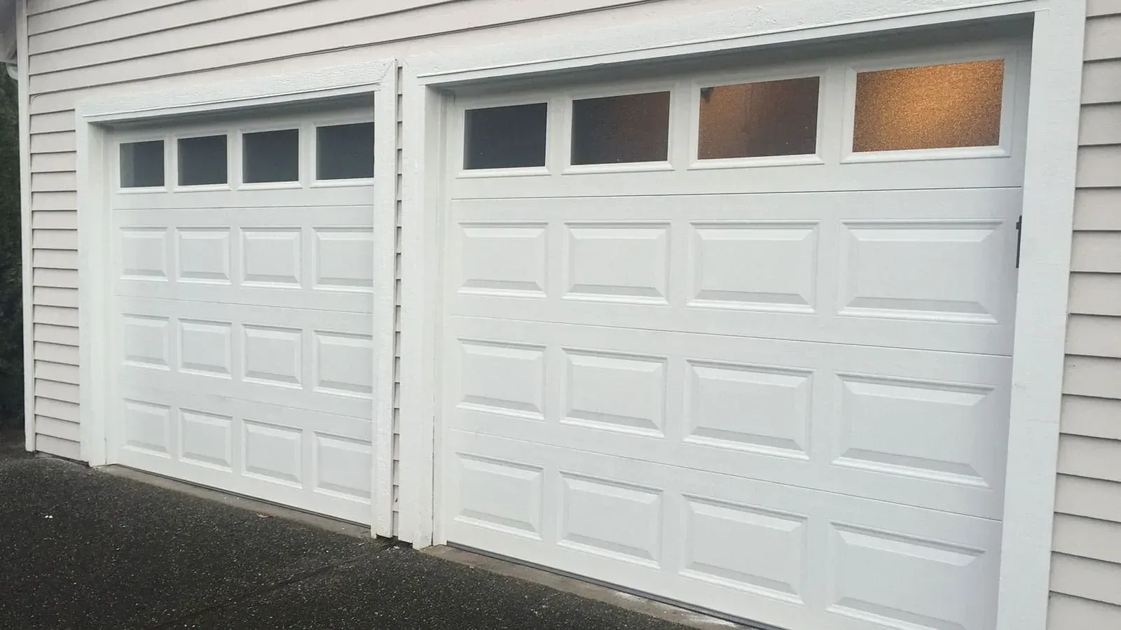 Two white garage doors with rectangular windows on top, installed side-by-side on a house with light-colored siding.