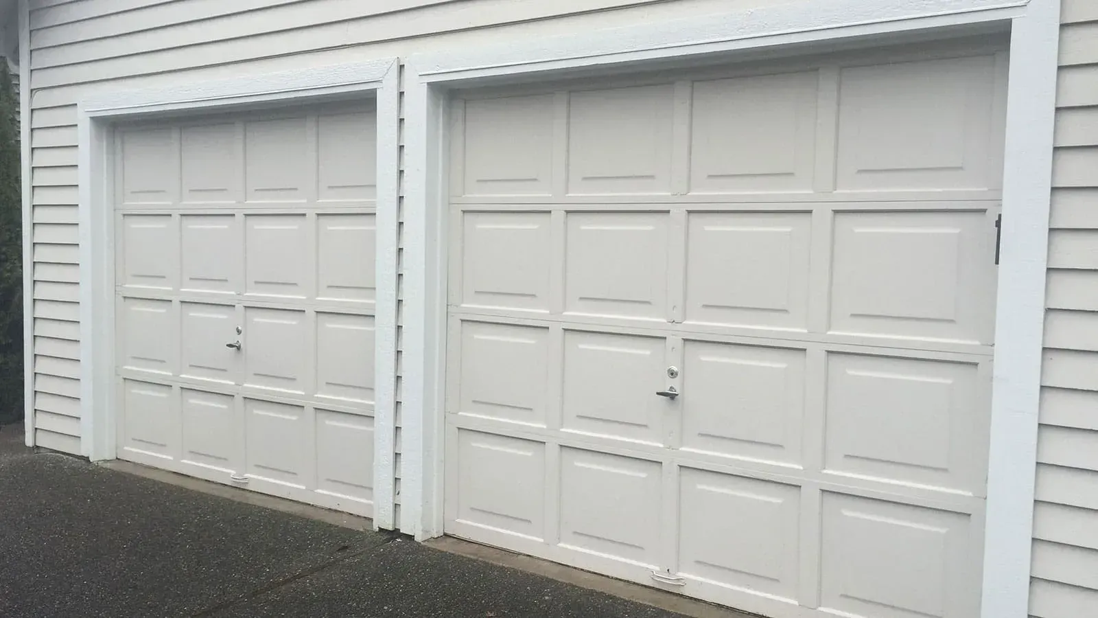 Two adjacent, light-colored rectangular garage doors with a grid panel design, set against white horizontal siding.