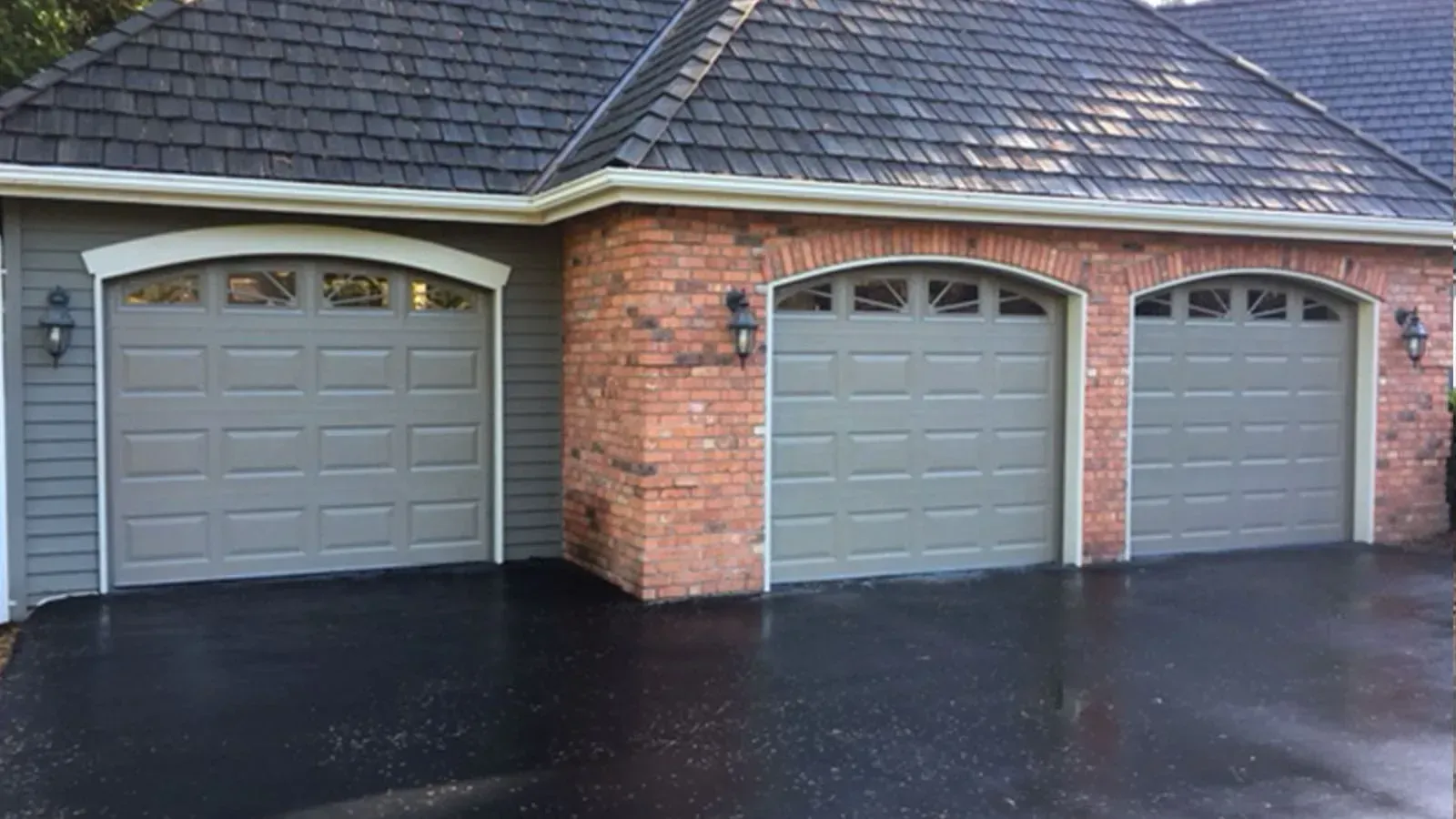 Three gray garage doors with arched windows set in a brick and siding home exterior with a dark asphalt driveway.