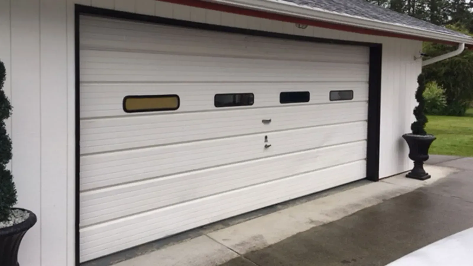 A white garage door with four rectangular windows, framed by black trim on a building exterior with potted topiary trees.